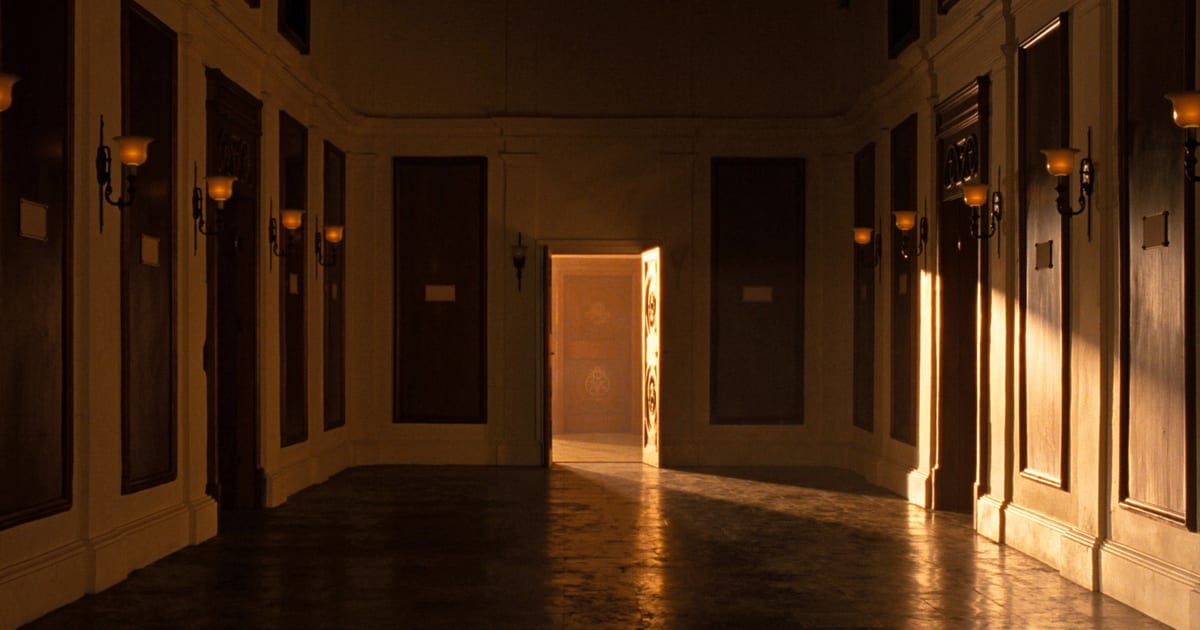 A dim panelled hallway lined with identical closed doors, one standing open onto bright light.